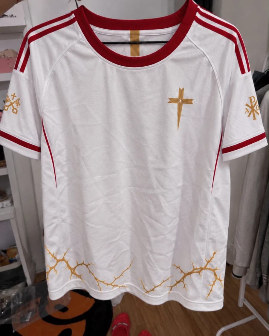 Le maillot sur un cintre, face visible : col et bandes rouges, croix dorée côté cœur.