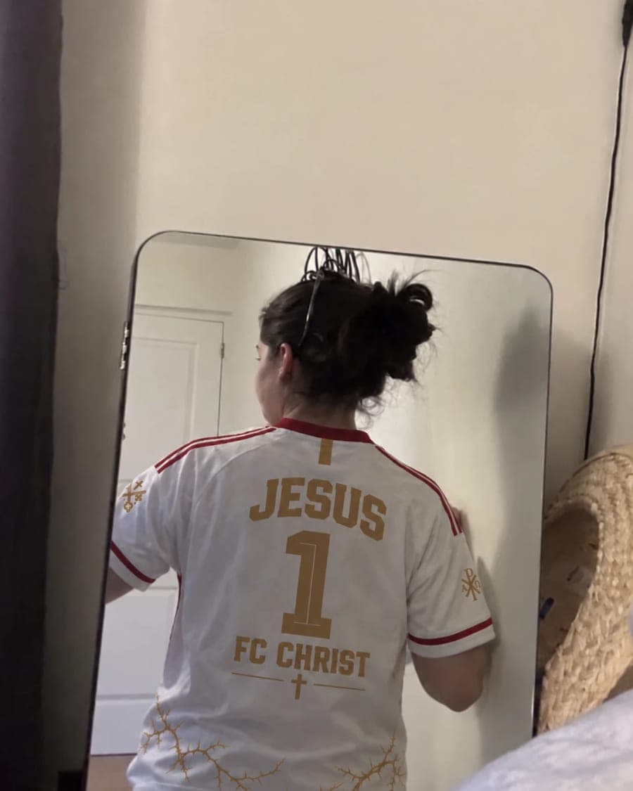 Le maillot porté de dos devant un grand miroir, flocage « JESUS 1 — FC CHRIST » en or.